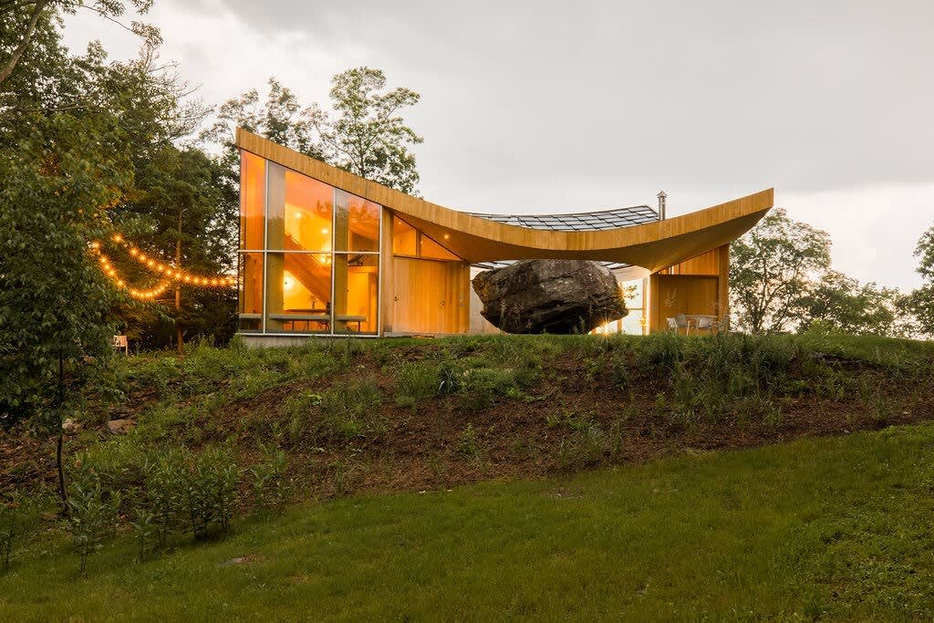 The distinctive house, with its curved wooden roof and large glass facade, is nestled on a grassy hillside surrounded by lush trees under a dramatic cloudy sky. String lights add a festive touch to the outdoor area, highlighting the unique integration with nature.