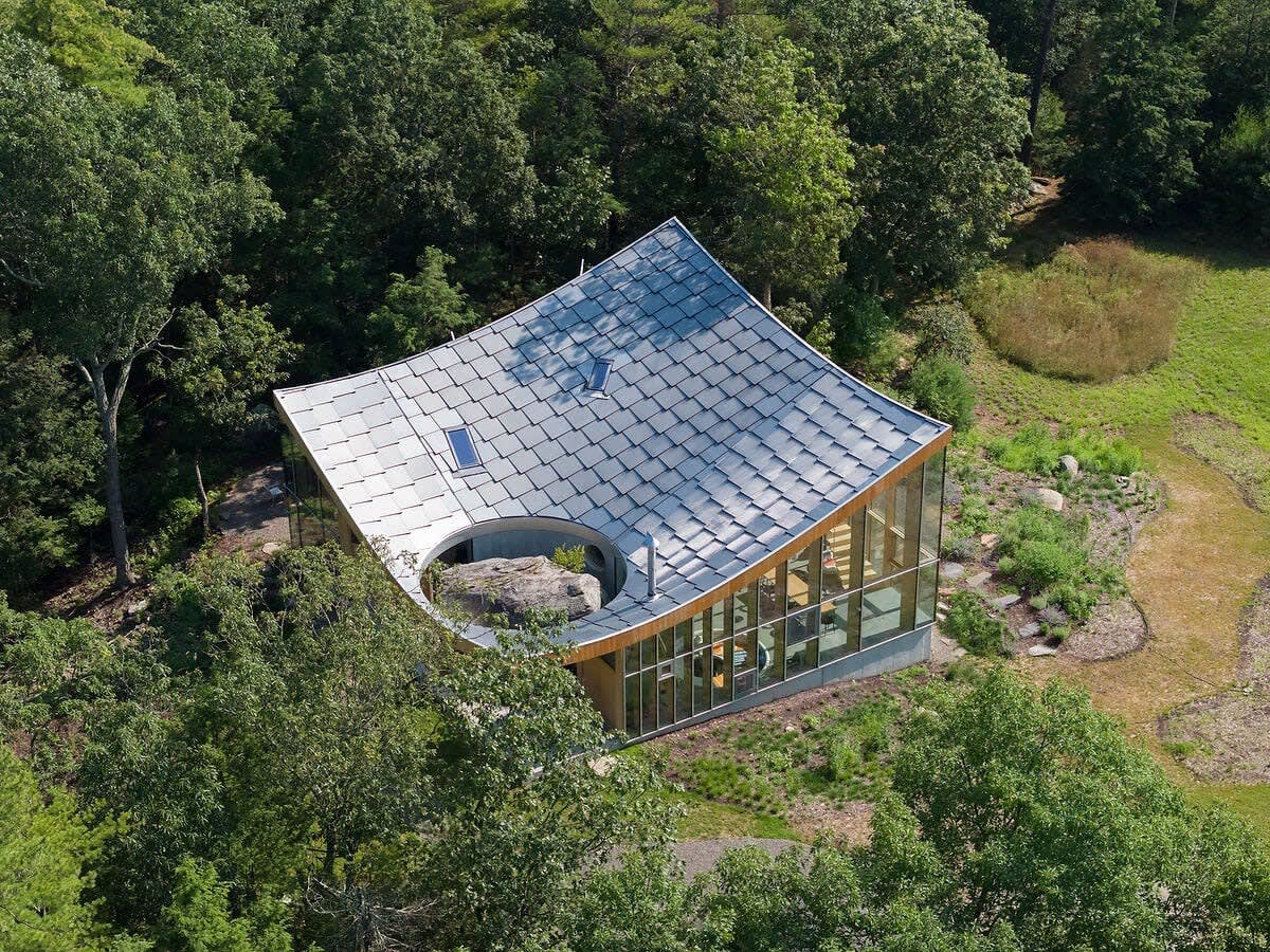 An aerial view showcases the distinctive curved, shingled roof of the house, with a circular cutout revealing a massive natural rock feature. The modern structure is surrounded by vibrant green trees and a meticulously landscaped yard.