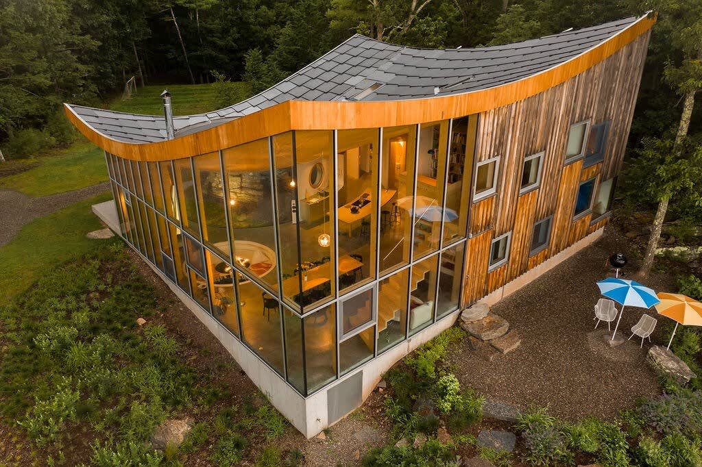An aerial view showcases the house's distinctive curved roof and extensive glass walls, revealing the open-plan interior bathed in light. The structure is surrounded by a mix of gravel and grass, nestled among dense green trees, highlighting its unique integration with the natural landscape.