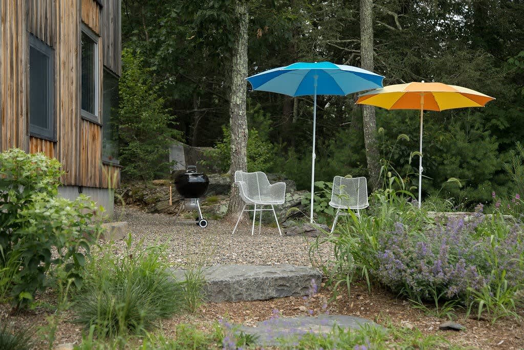 A cozy outdoor patio area features a black BBQ grill, two white mesh chairs, and two colorful umbrellas (blue and orange) on a gravel ground, surrounded by lush green plants and a wooden-sided house.