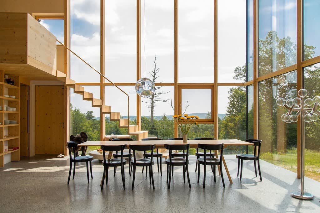 Spacious dining area with a long wooden table and black chairs, positioned in front of a dramatic wall of floor-to-ceiling windows that showcase a distant mountain and forest landscape. A modern staircase with wooden steps is visible to the left.
