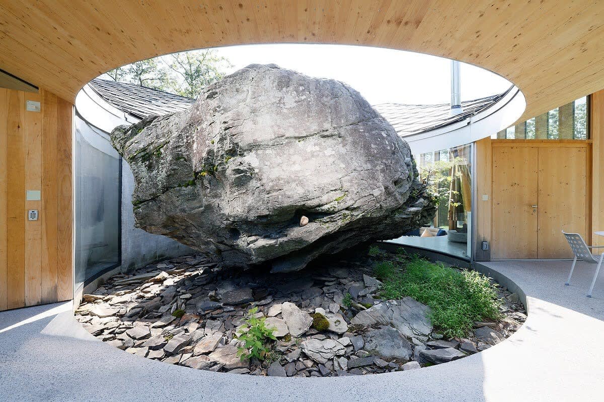 A massive natural rock dominates a circular open-air courtyard, surrounded by smaller rocks and sparse greenery, framed by the house's wooden walls and a unique roof opening.