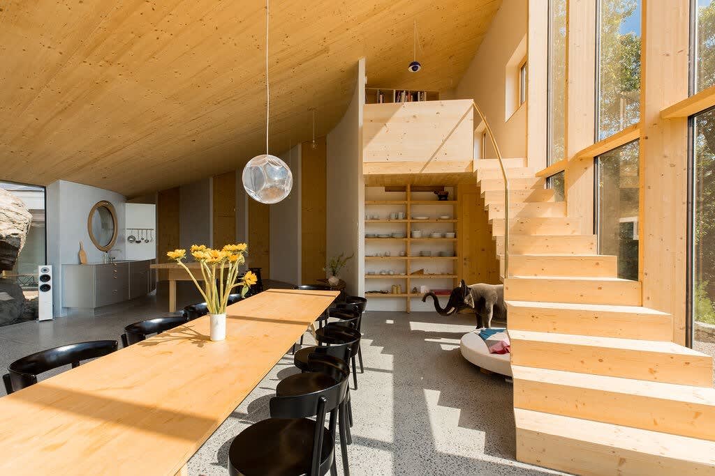 Wide view of the open-plan living and dining area, showcasing a long wooden dining table with black chairs, a modern wooden staircase, and a glimpse of the kitchen area with a circular window. The space is characterized by high wooden ceilings and large windows.