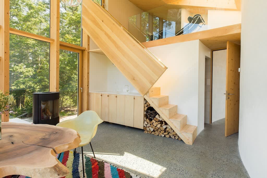 A bright common area showcases a unique wooden staircase with integrated firewood storage underneath, leading to an upper level. A light wood counter and cabinets are built into the wall, complemented by large windows offering outdoor views and a concrete floor.