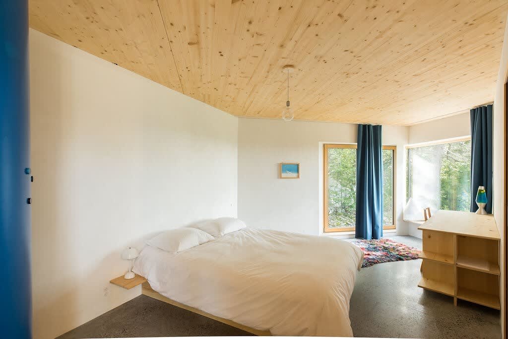 A serene bedroom with a white-covered bed, light wood ceiling, and large windows offering a view of greenery. A small wooden side table and a colorful rug add warmth to the space.
