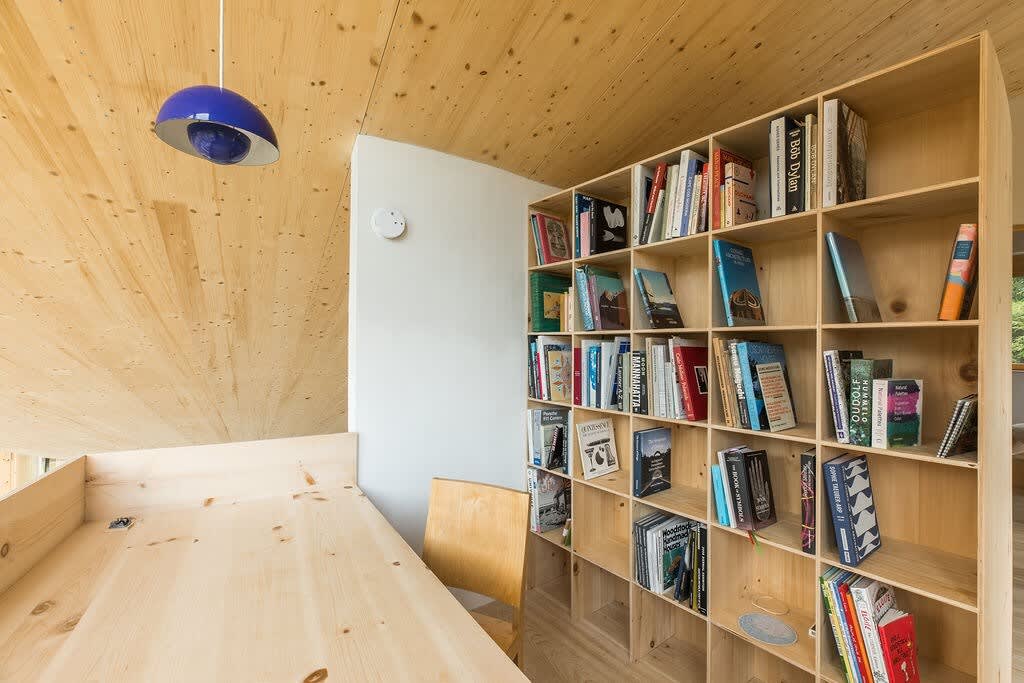 A cozy office nook with a light wood sloped ceiling and a large built-in wooden bookshelf filled with books. A simple wooden desk and chair are visible, illuminated by natural light.