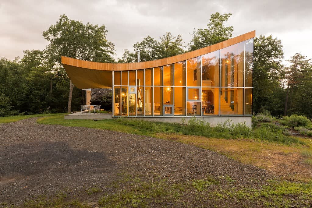 A striking modern house with a curved wooden roof and a full glass front is set against a backdrop of green trees and a cloudy sky. A gravel driveway leads to the entrance, and a small patio with outdoor seating is visible on the side.