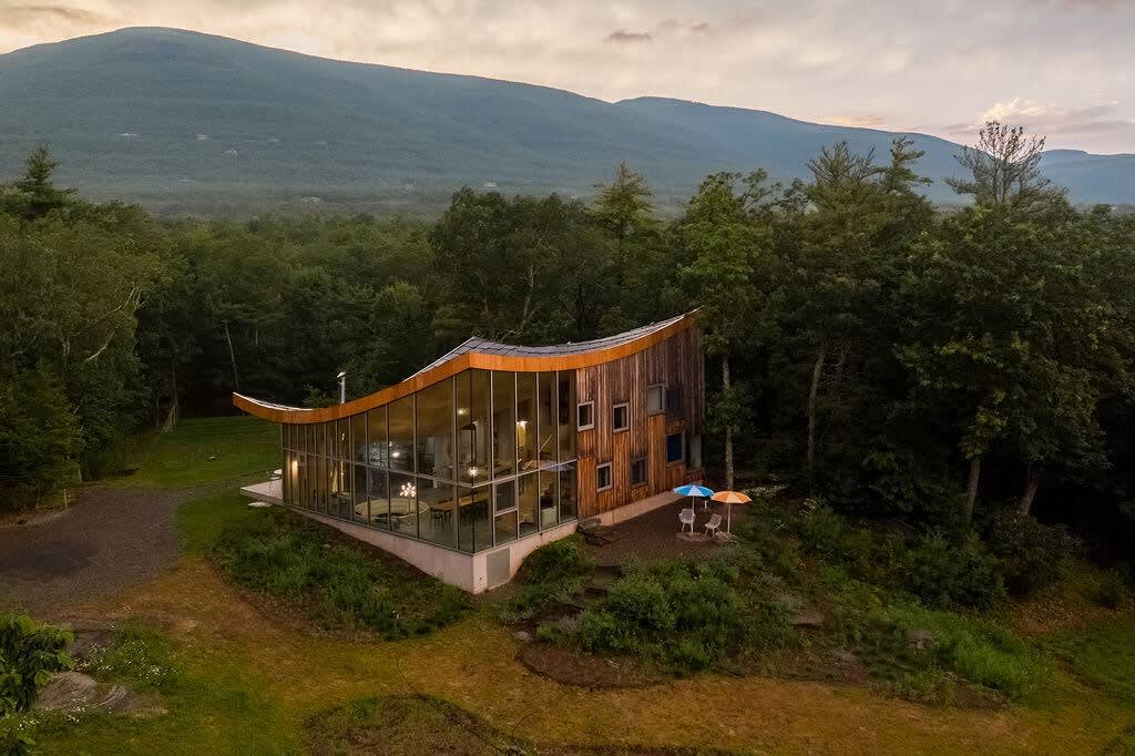 An aerial-like view captures the unique curved-roof house with a glass front, nestled in a clearing surrounded by dense forest and distant mountains. The property features a grassy lawn and a small outdoor seating area, blending seamlessly with nature.