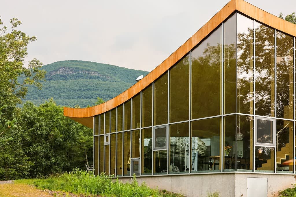 The modern house boasts a distinctive curved wooden roof and floor-to-ceiling glass walls, reflecting the surrounding lush greenery, with a verdant mountain visible in the background.