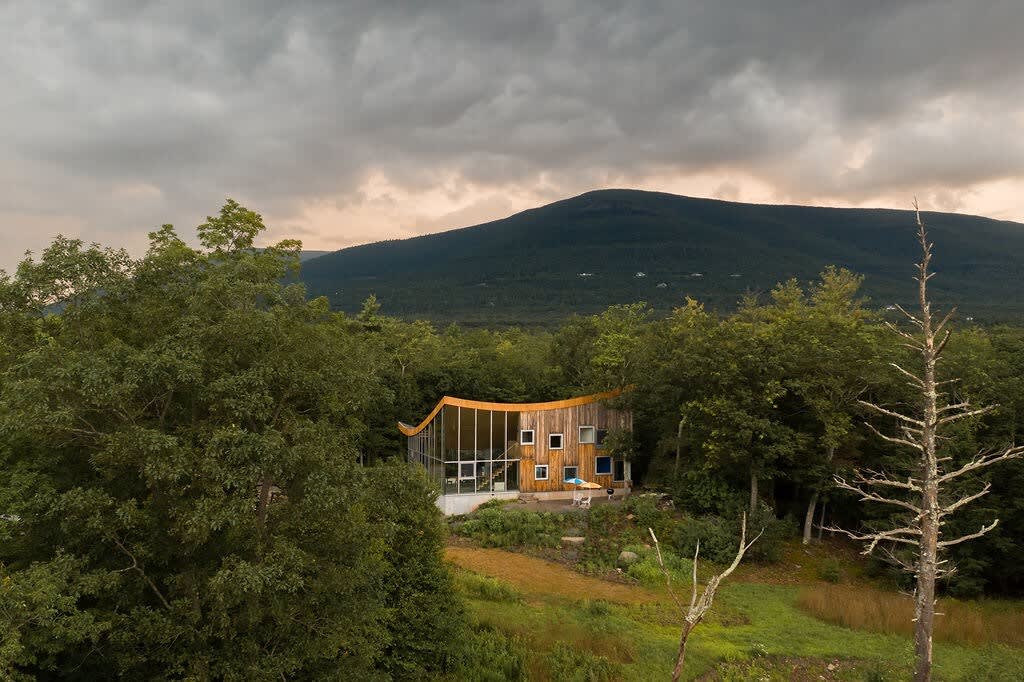 The unique house, with its curved roof and glass facade, is nestled into a lush, tree-covered mountainside under a dramatic cloudy sky. The natural setting emphasizes the property's secluded and scenic location, blending modern architecture with the rural landscape.