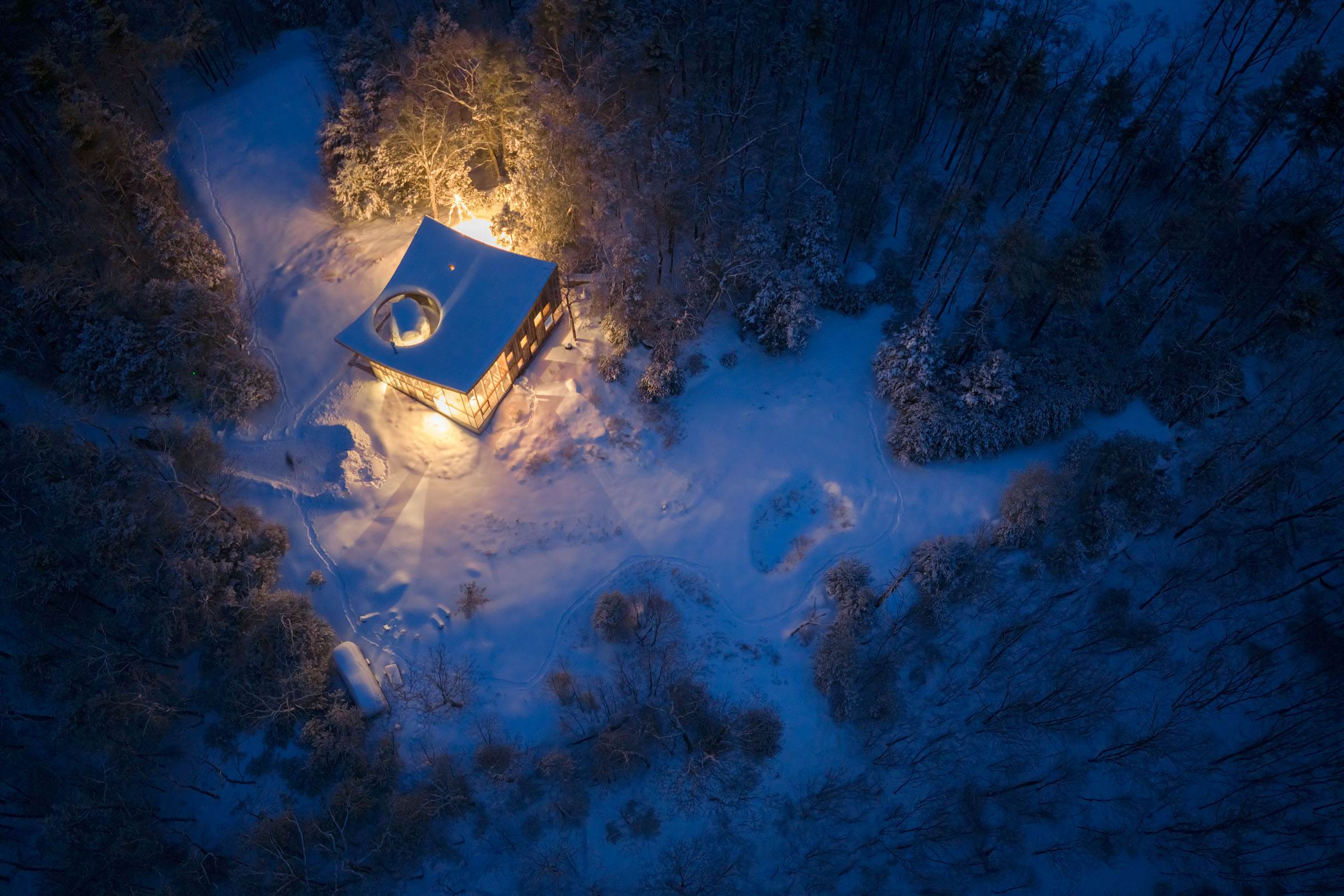 A closer aerial night shot highlights the illuminated house, its unique curved roof visible, set against a pristine snow-covered landscape with visible paths winding through the surrounding winter forest.