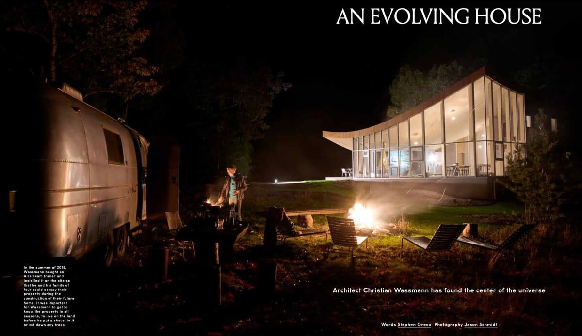 The modern house glows warmly at night, showcasing its curved glass facade, while an Airstream trailer and a crackling fire pit with Adirondack chairs create a rustic outdoor gathering space in the foreground.