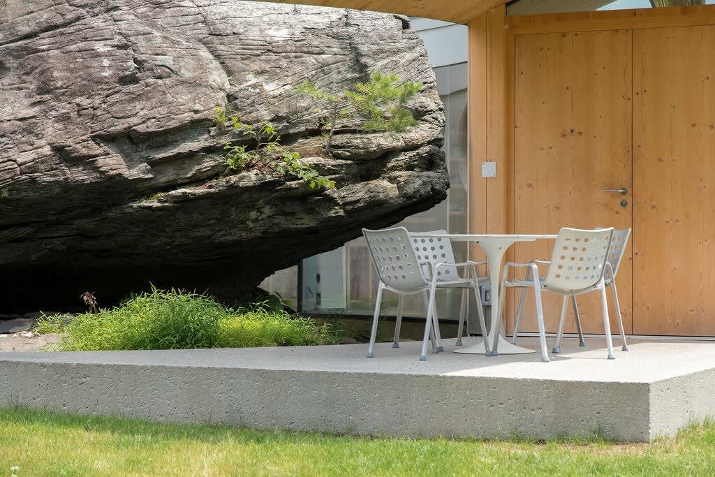 An inviting outdoor patio area features a white table and chairs, perfectly situated next to a massive natural rock formation that is seamlessly integrated into the house's design. Green foliage surrounds the concrete patio, enhancing the natural ambiance.