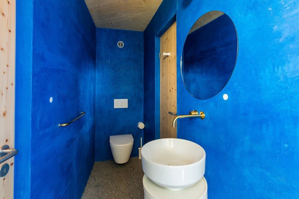 A bright blue bathroom featuring a wall-mounted toilet, a white vessel sink with a brass wall-mounted faucet, and a large round mirror. The room has a natural wood door and ceiling.