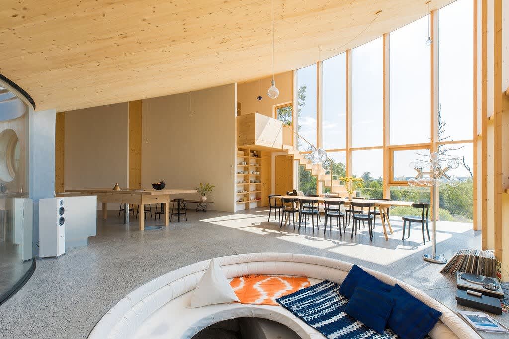 Vast open-plan living area with a high wooden ceiling and polished concrete floors, featuring a sunken circular seating area in the foreground and a dining space with a long wooden table and chairs in the background, all bathed in natural light from expansive windows.