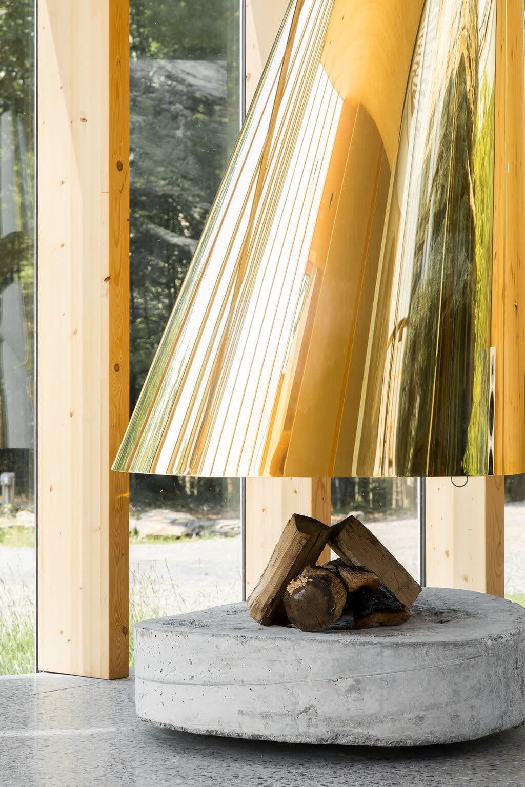 Close-up of the modern golden fireplace with a concrete base, featuring stacked firewood. The large windows behind it reveal lush green trees, emphasizing the connection to nature.