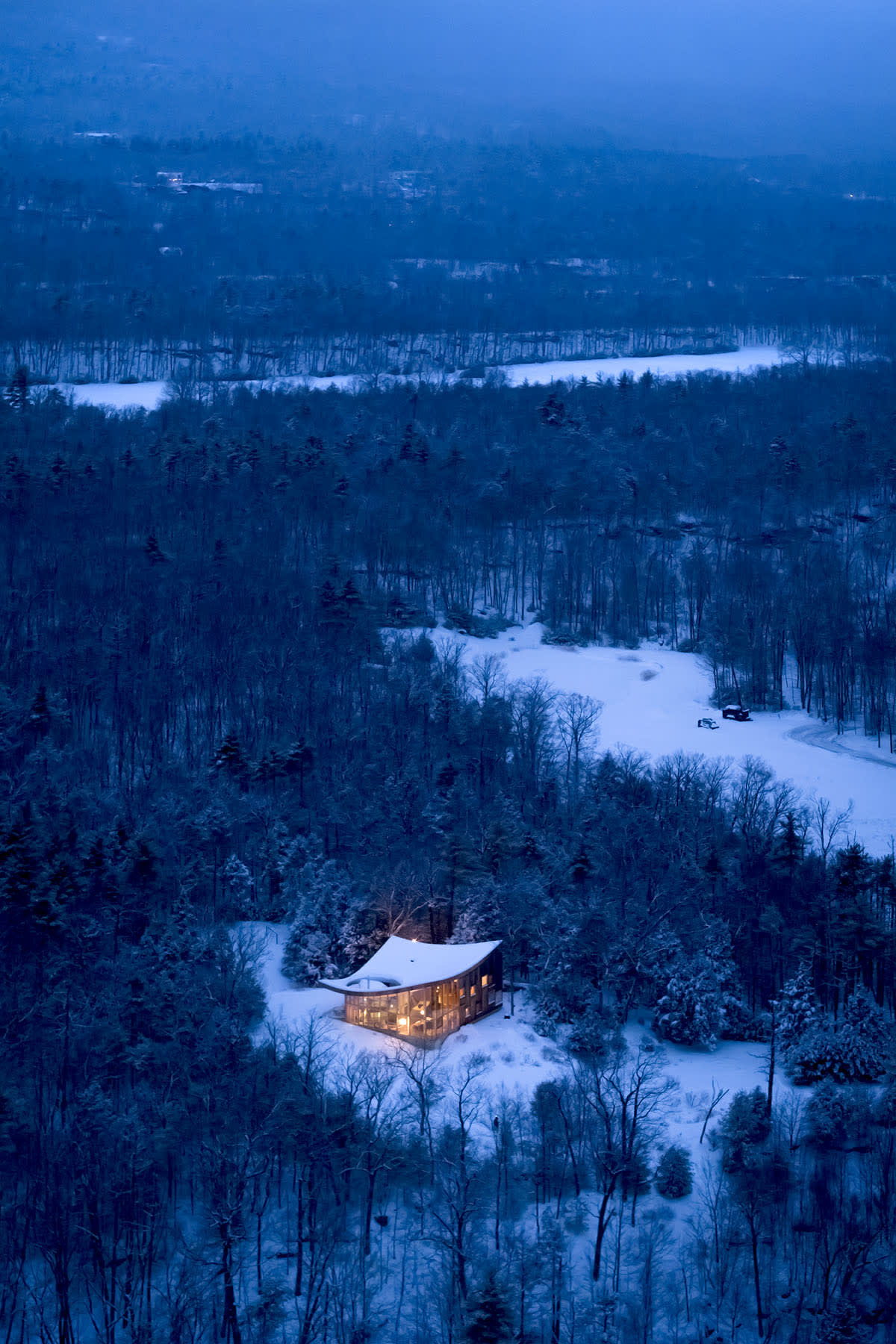 An aerial view of a modern, curved-roof house nestled in a snow-covered forest at dusk, with warm lights glowing from its large windows, creating a cozy contrast against the blue winter landscape.