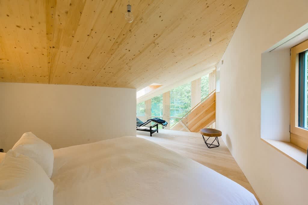 A spacious bedroom features a large white bed, light wood flooring, and a sloped wood-paneled ceiling, creating an airy atmosphere. A window offers a glimpse of the surrounding trees, and a black lounge chair with a small stool is visible in the background.