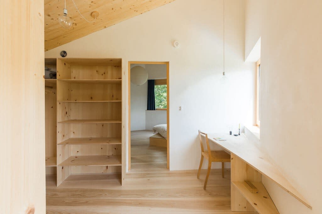 A bright, minimalist interior space with light wood floors and ceiling, featuring built-in wooden shelving and a long wooden desk extending from the wall. A doorway leads to another room with a bed visible.
