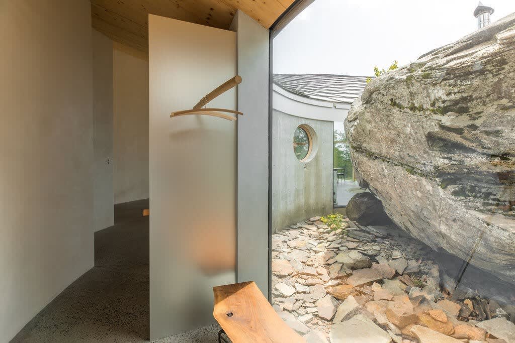 A minimalist interior hallway features light-colored walls and a wooden coat rack, with a large window offering a direct view of the massive natural rock outside. The concrete floor and simple design highlight the connection between the interior and the unique exterior landscape.
