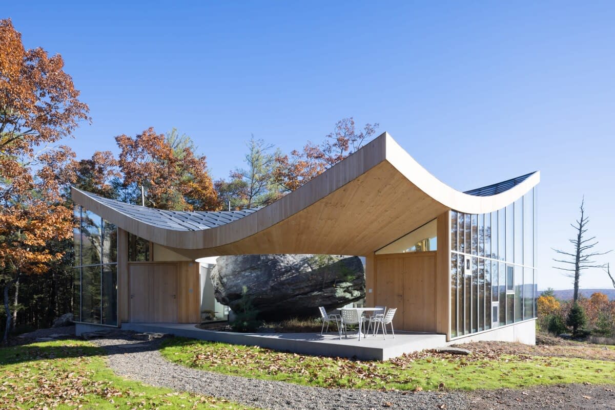 The unique architectural exterior of the house features a dramatic curved wooden roof and extensive glass walls, framing a massive natural rock at its core. A small outdoor patio with seating is nestled beside the structure, surrounded by autumn foliage and a gravel path.