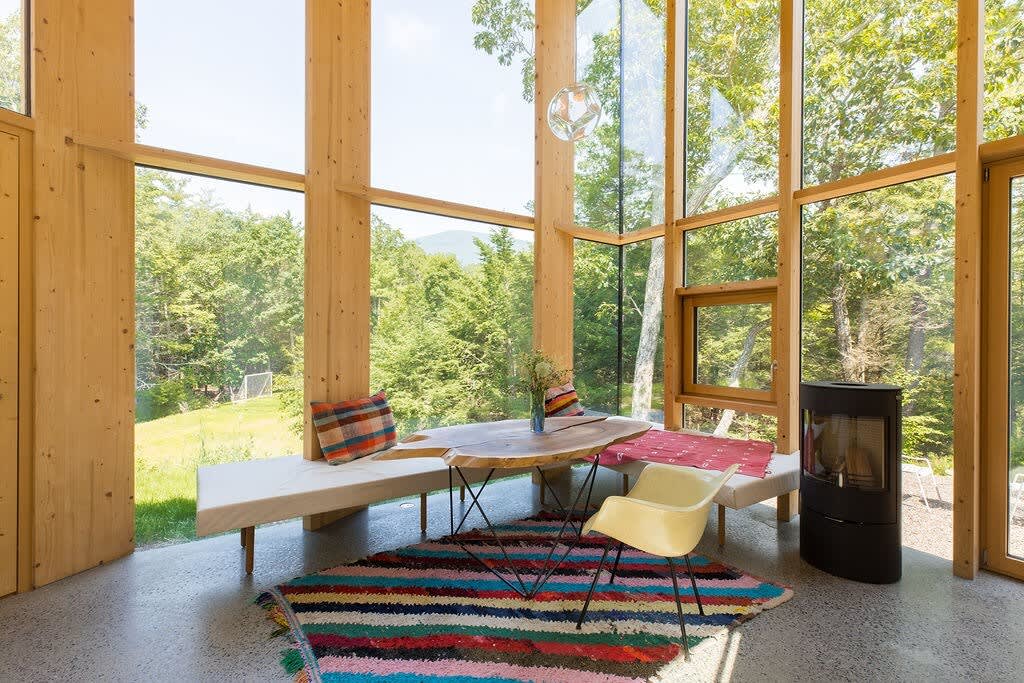 A cozy lounge area features a built-in bench with cushions, a unique live-edge wooden table, and a colorful striped rug. Tall windows provide abundant natural light and views of the surrounding forest, complemented by a sleek black wood-burning stove.