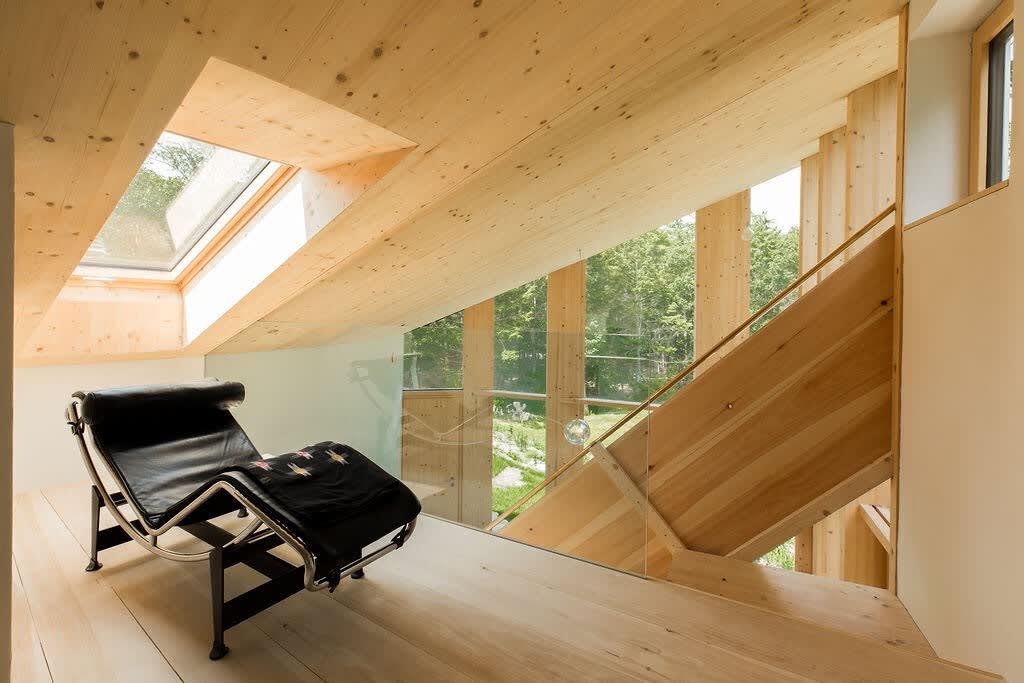 A bright loft area with light wood flooring and a sloped wood ceiling features a black leather lounge chair, perfect for relaxation. A glass railing overlooks the lower level, and expansive windows provide stunning views of the surrounding forest.