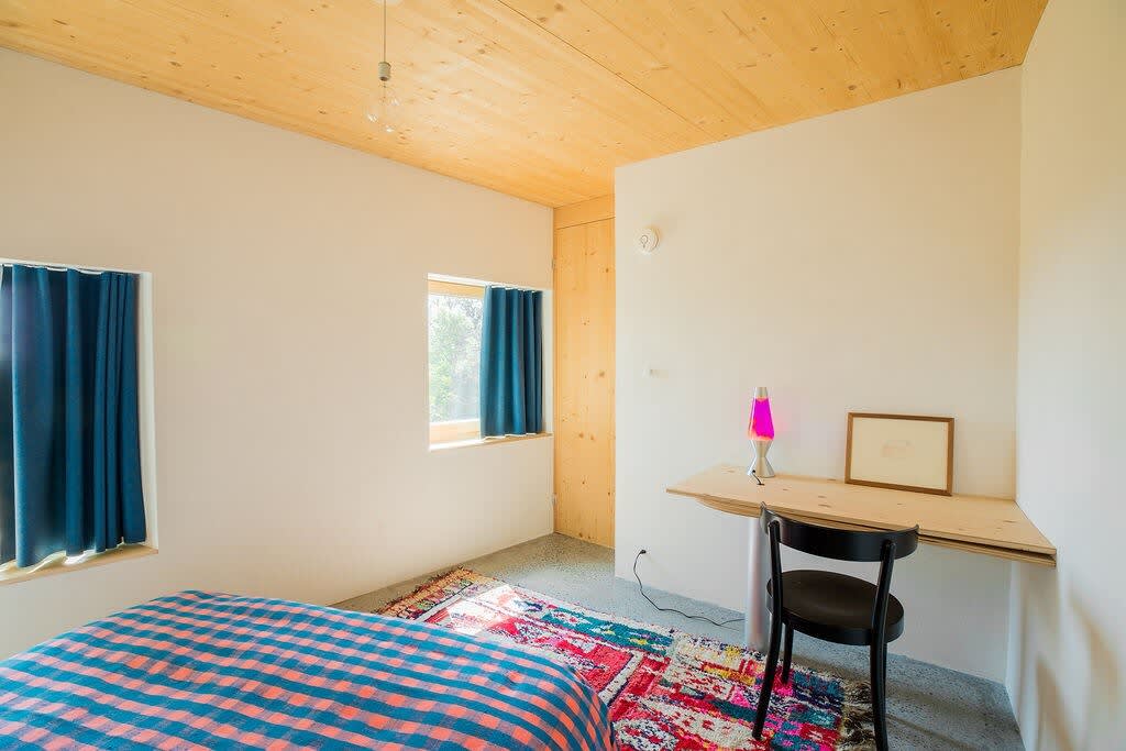 A cozy bedroom features a bed with a blue and orange plaid comforter, a light wood ceiling, and a small desk with a lava lamp. Two windows with blue curtains provide natural light, illuminating the area rug and concrete floor.