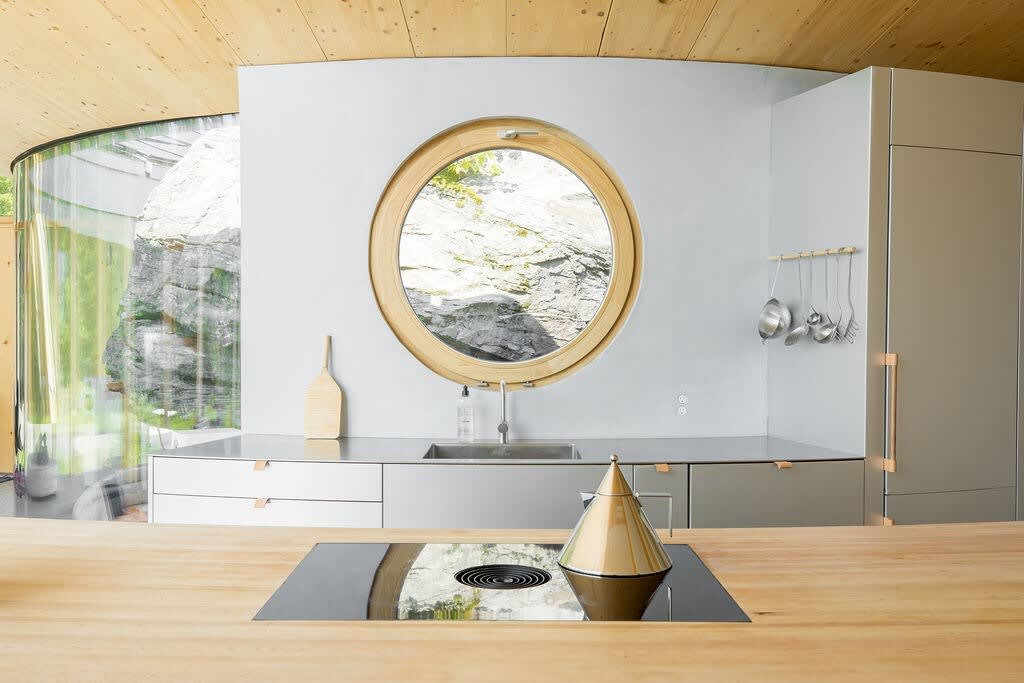 Contemporary kitchen with light gray cabinetry, a stainless steel sink, and a unique circular porthole window framing a view of a rocky outcrop. A wooden countertop with an integrated cooktop is in the foreground.