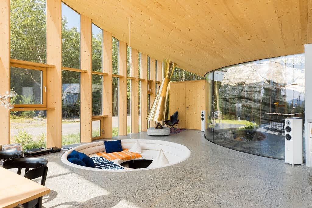 Open-concept living space featuring a unique sunken circular seating area with colorful cushions, a striking golden fireplace, and a curved glass wall offering views of a rocky outdoor area.