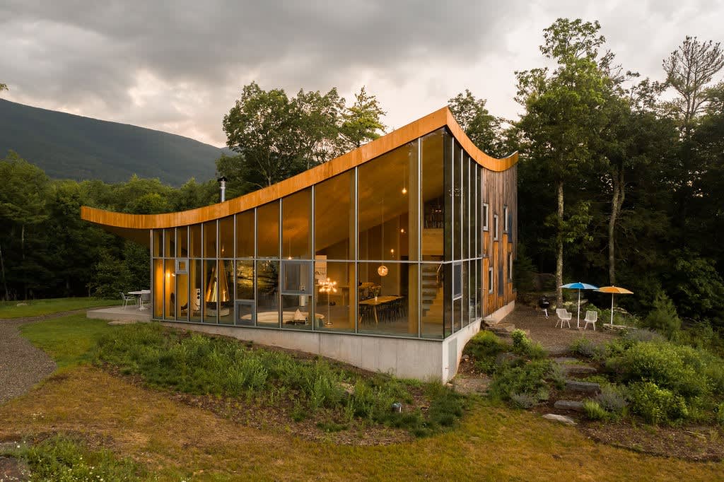 An architectural house with a distinctive curved roof and a glass wall facade is situated on a grassy slope surrounded by lush trees. Distant mountains are visible under a cloudy sky, highlighting its serene rural setting.