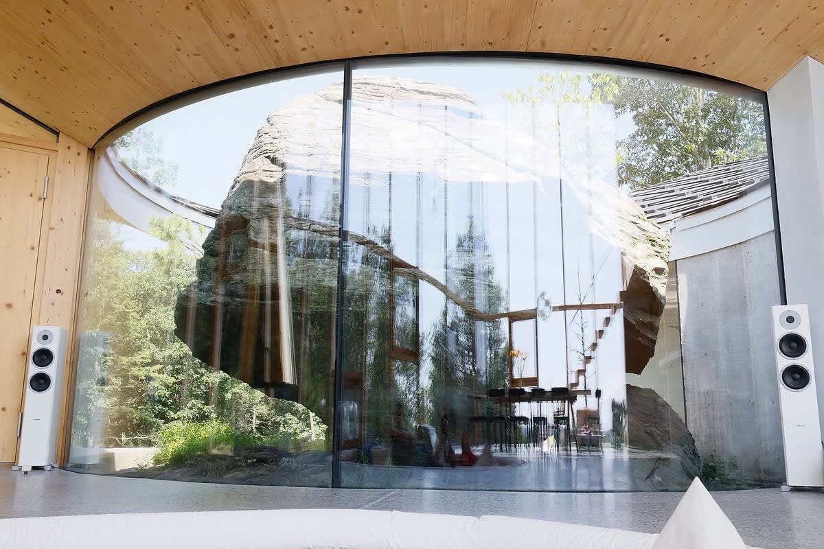 A spacious living area features a dramatic curved glass wall offering an immersive view of a massive natural rock formation, complemented by a light wooden ceiling and modern white speakers.