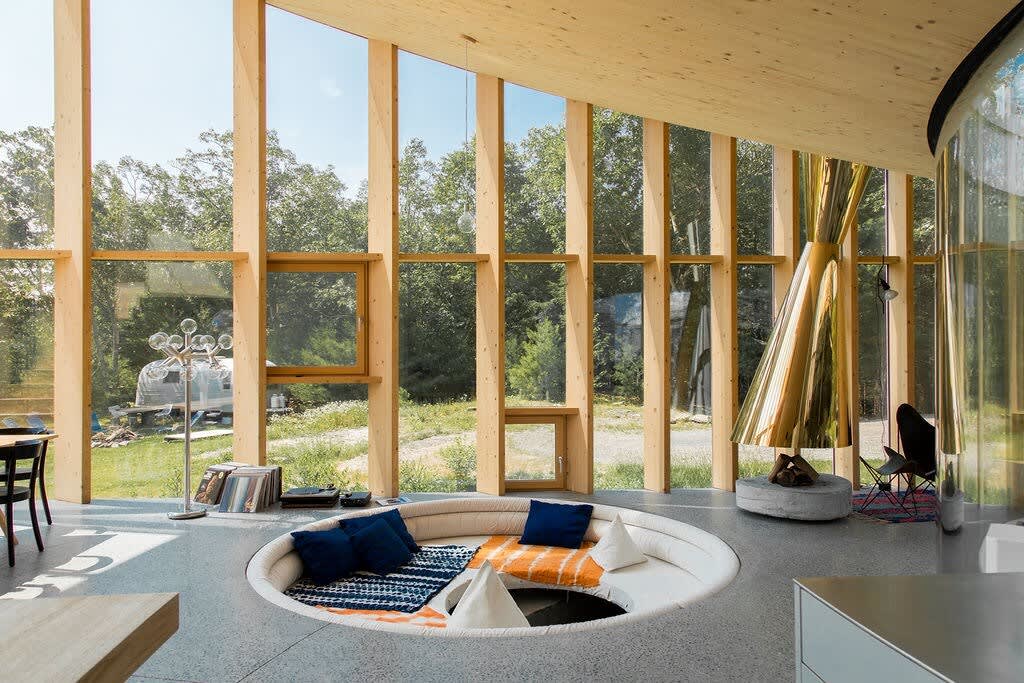 A modern living room features a circular sunken seating area with blue and orange cushions, centered around a unique golden fireplace. Expansive floor-to-ceiling windows offer breathtaking views of the surrounding forest, all beneath a curved wooden ceiling.