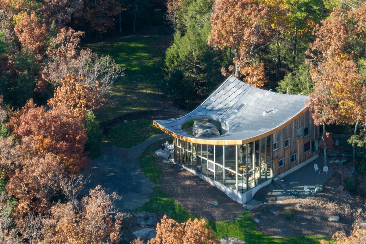 An aerial view showcases the unique, curved-roof house nestled among trees with vibrant autumn foliage, featuring extensive glass walls and a central open-air rock formation.