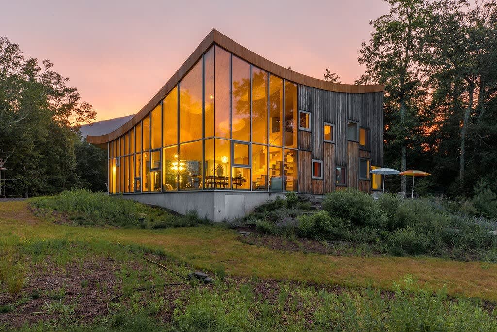 The unique, modern house with a curved roofline and extensive glass facade glows warmly at dusk, nestled among trees. The wooden side wall features various small windows, and the sky shows beautiful sunset colors.