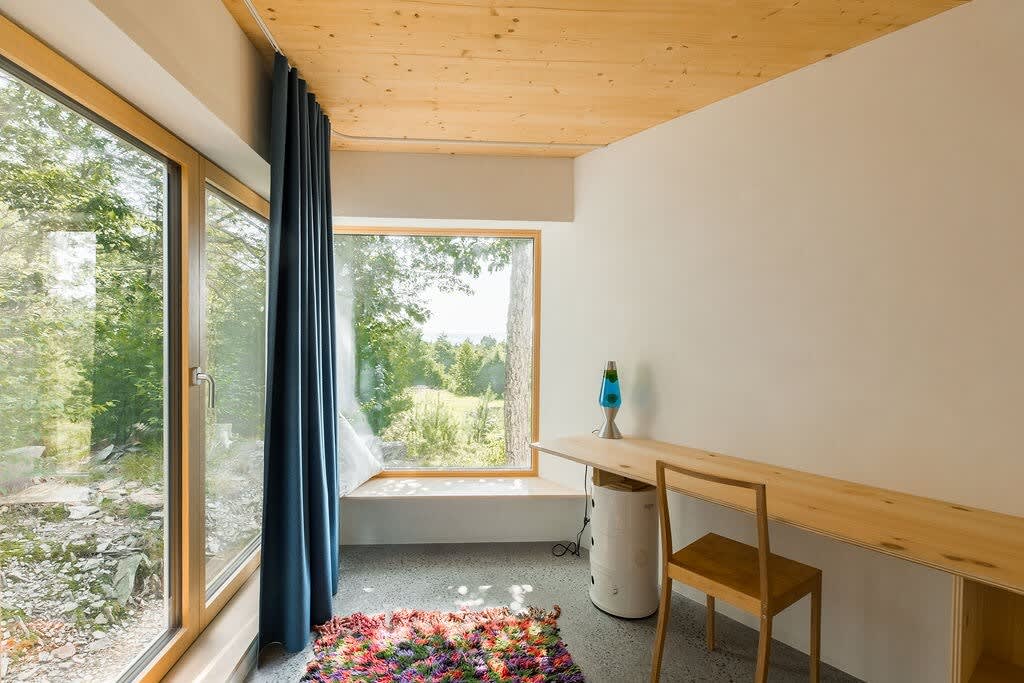A bright corner of a bedroom featuring a long wooden desk built into the wall, a simple wooden chair, and expansive windows overlooking a lush green landscape. A colorful rug and a lava lamp add character.