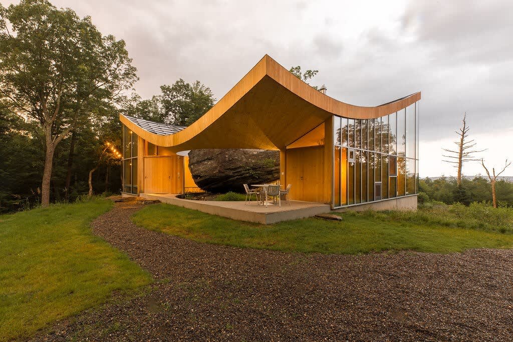 The unique house features a dramatic curved wooden roof and expansive glass walls, with a massive natural rock integrated into its design, alongside an outdoor patio with a table and chairs. A gravel path leads to the entrance.