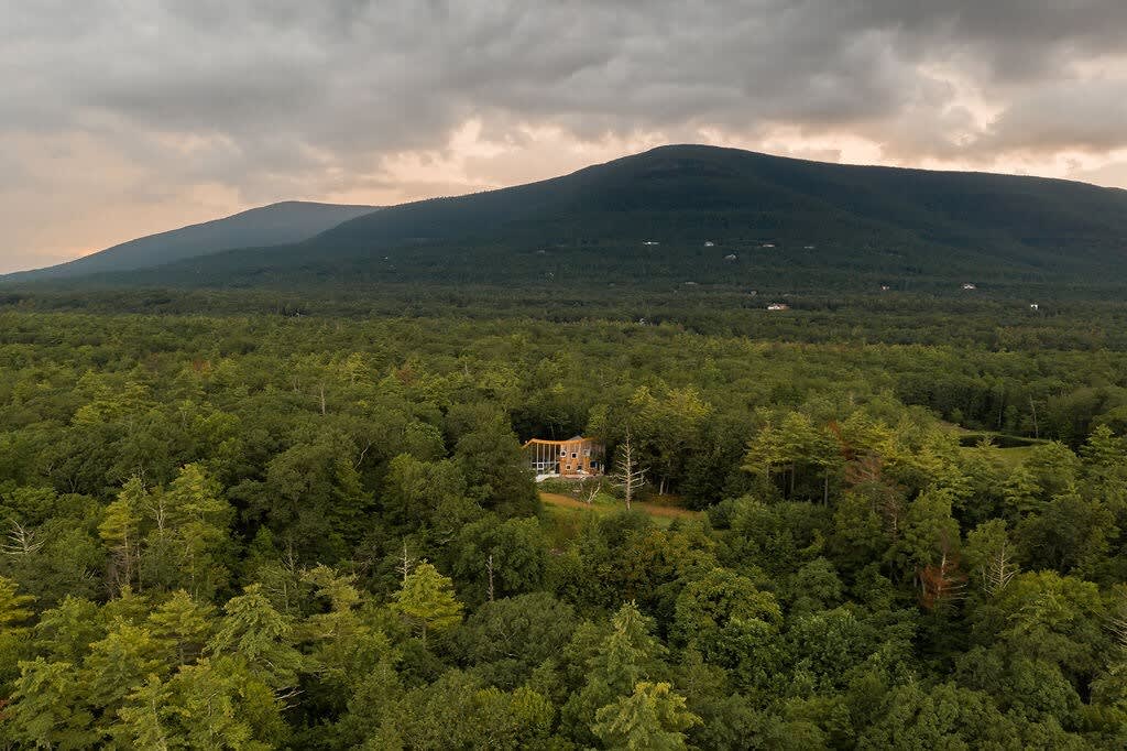 A distant aerial view reveals the unique house tucked away in a dense green forest, with majestic mountains rising in the background under a cloudy sky.
