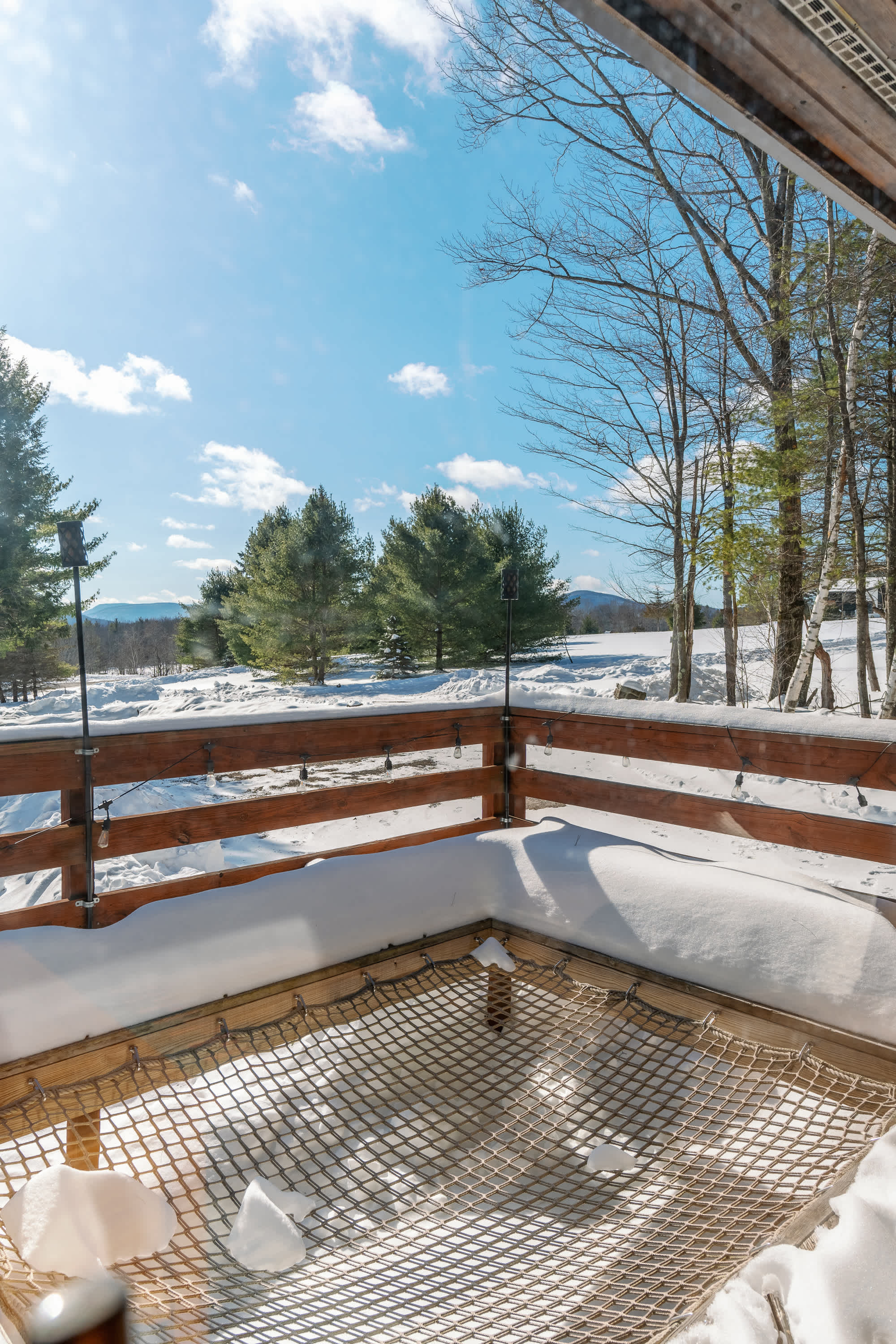 A snow-covered wooden deck features a built-in net hammock, offering panoramic views of a winter landscape with evergreen trees and distant mountains under a bright blue sky. Wooden railings with string lights line the deck.