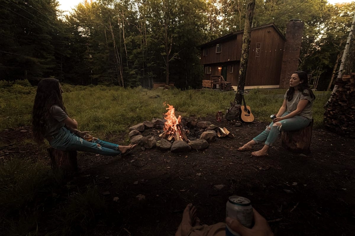 Two people sit on tree stumps around a crackling fire pit in a grassy clearing, with a rustic wooden cabin visible in the background surrounded by dense forest. A guitar and a can are also visible near the fire.
