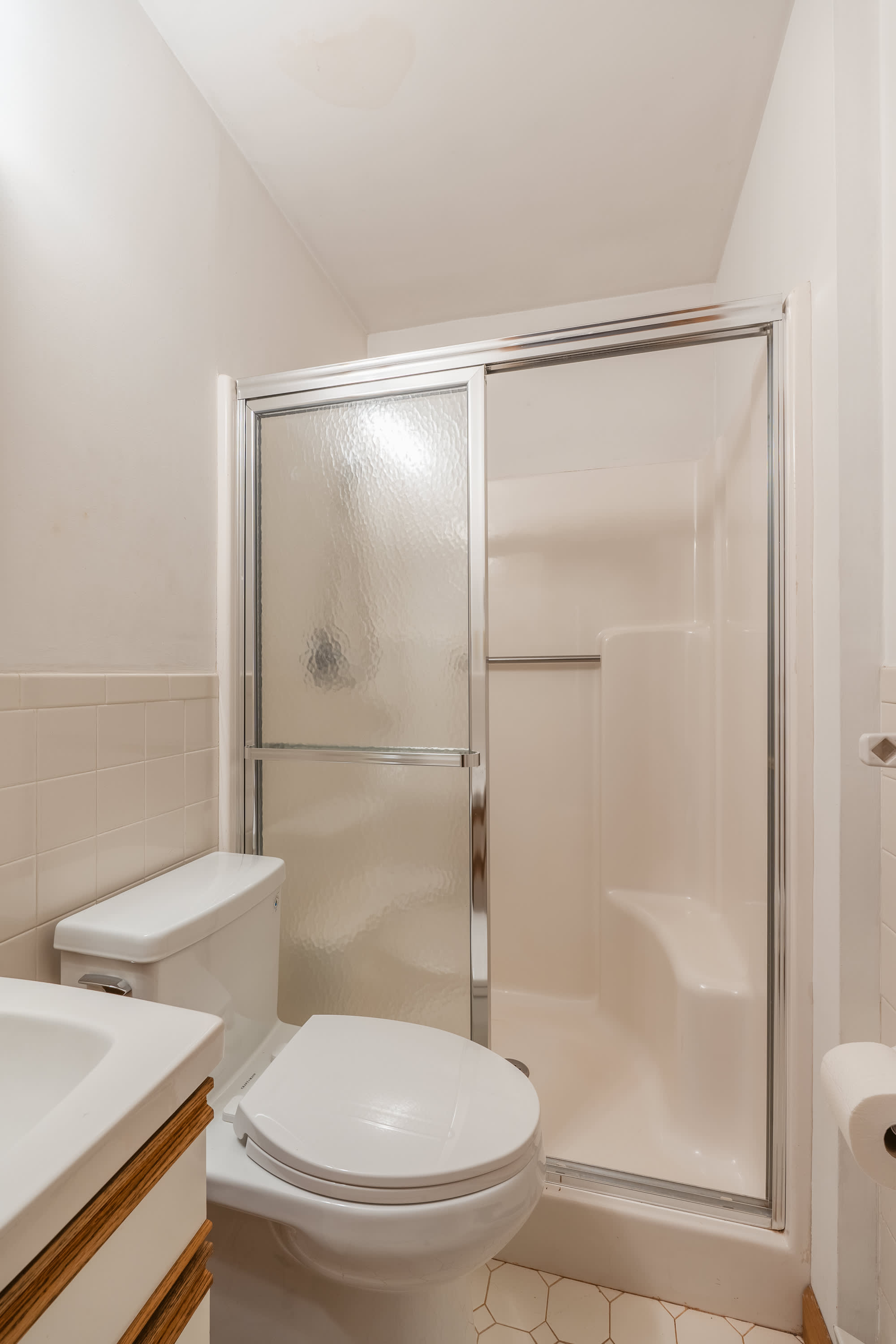A compact bathroom with a white toilet, a built-in shower stall with a sliding glass door, and light-colored tiled walls. A small sink vanity is partially visible on the left.
