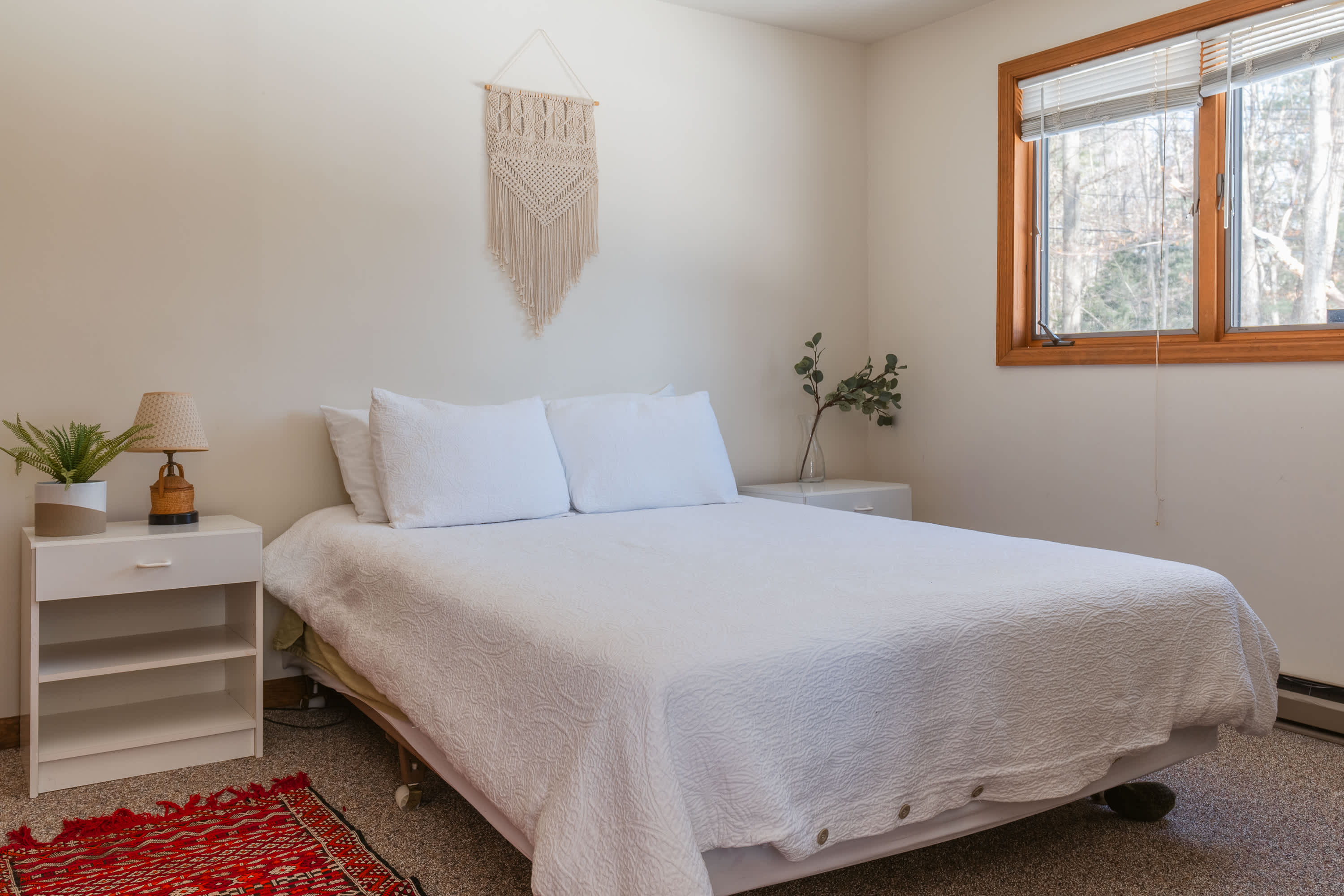A cozy bedroom featuring a queen-sized bed with white bedding, light-colored walls, and a macrame wall hanging above the bed. A wooden-framed window offers a view of trees, and a white nightstand with a lamp and plant sits beside the bed.