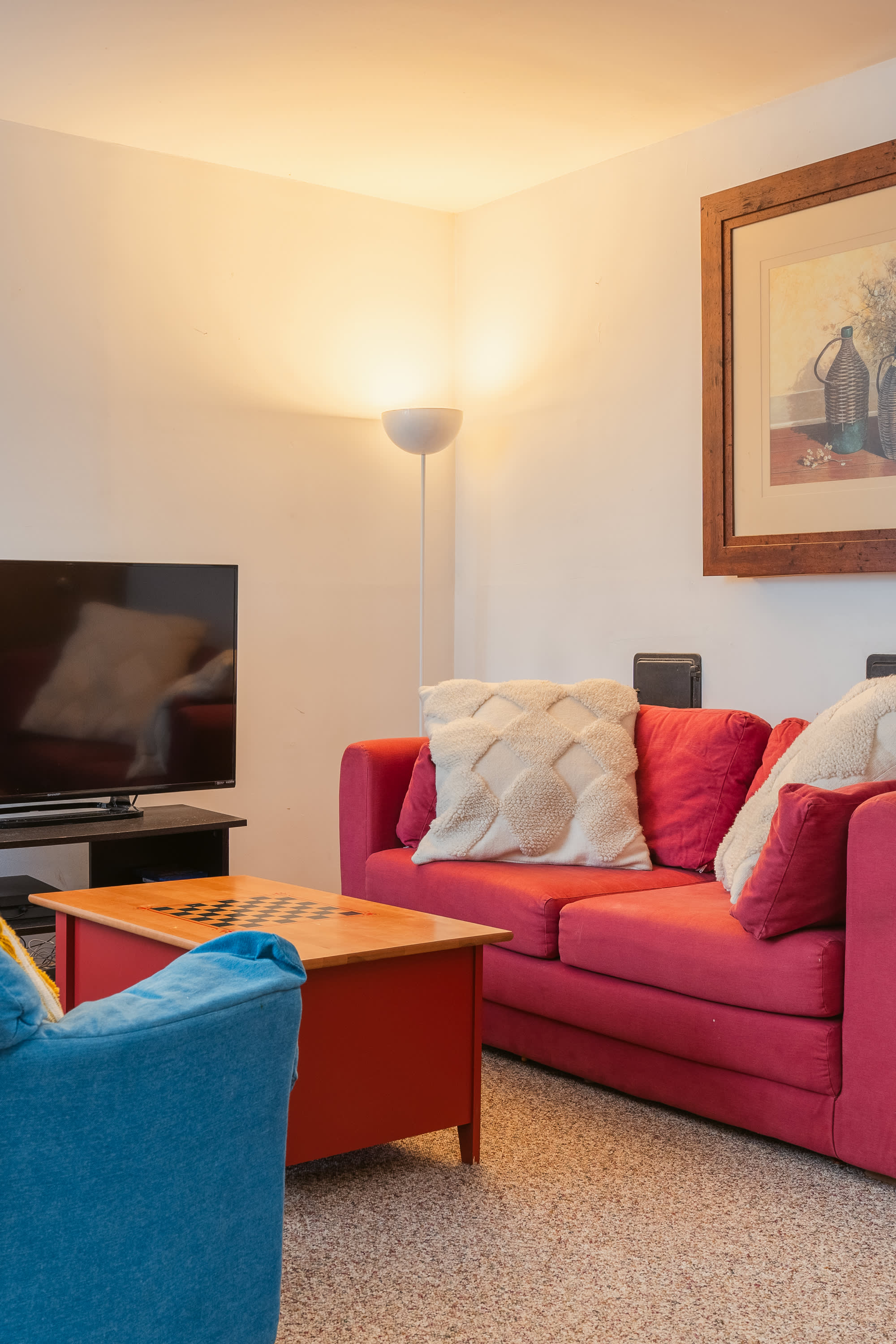 Cozy secondary living area with a red sofa, a blue armchair, and a coffee table with a chessboard. Features carpeted flooring, a floor lamp, and a wall-mounted TV.