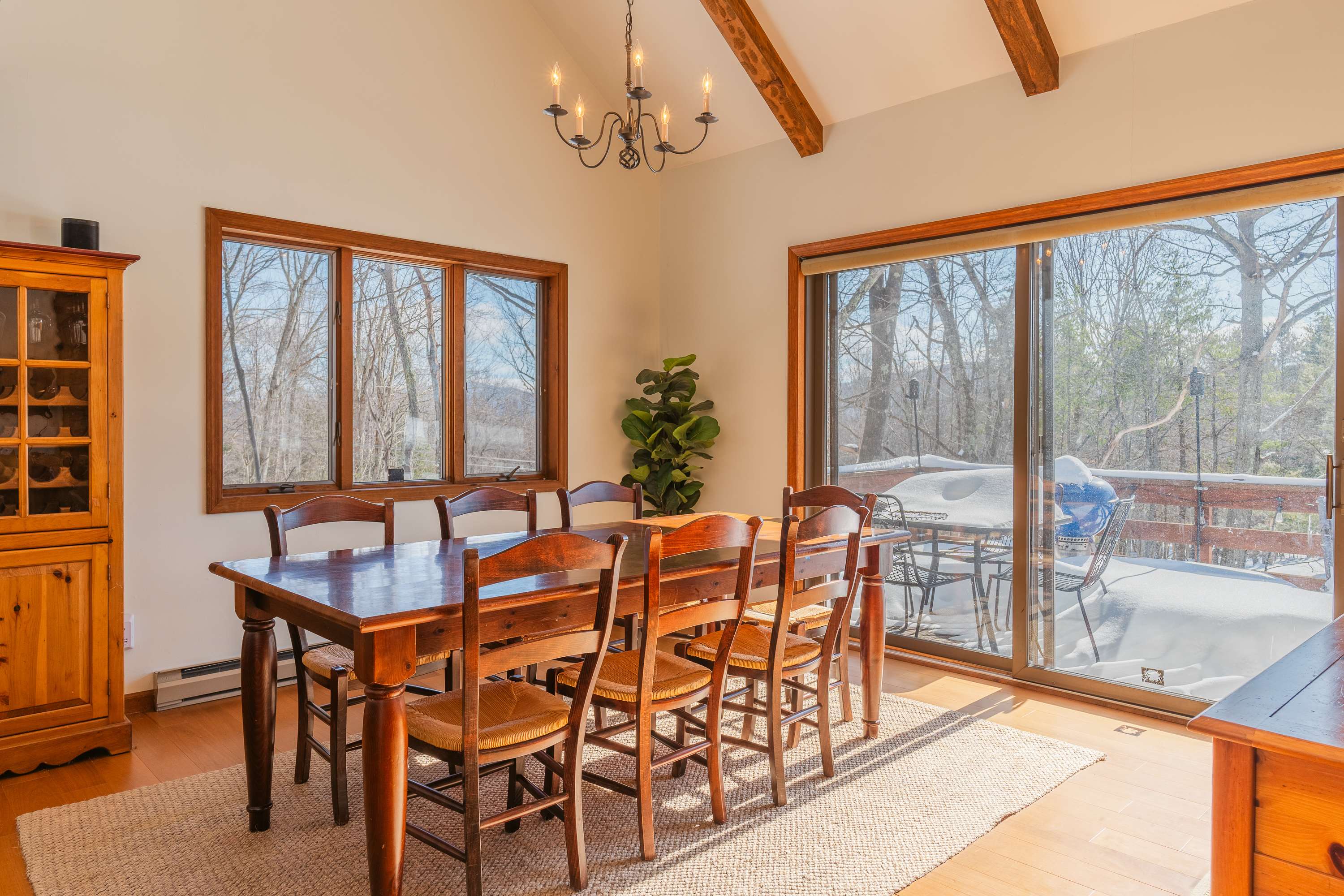 A sunlit dining area features a large wooden table with eight chairs, a wooden hutch, and sliding glass doors leading to a deck with a snowy outdoor view.