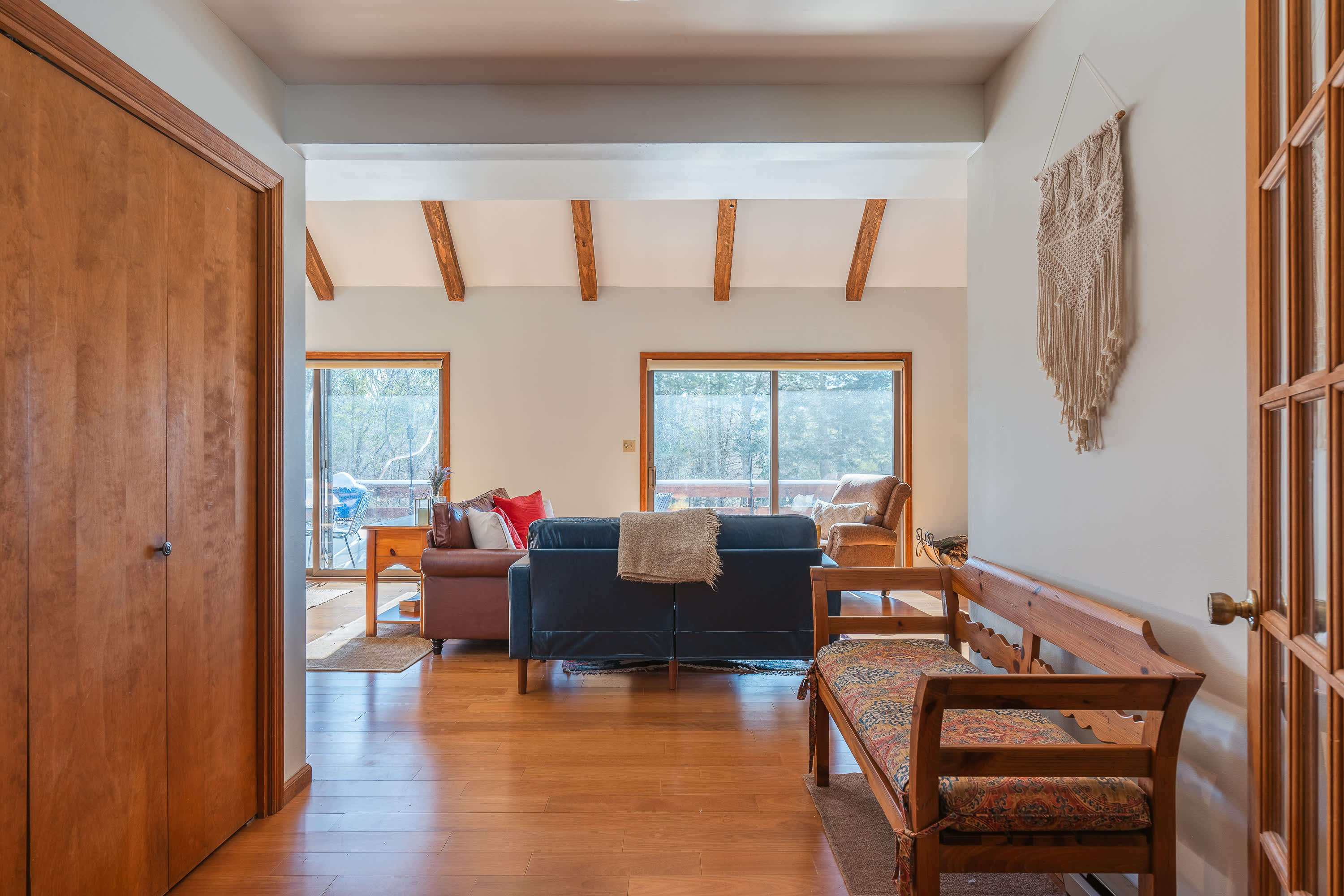 A bright entryway with hardwood floors features a wooden bench with a patterned cushion and a macrame wall hanging, leading into a spacious living area with large windows.
