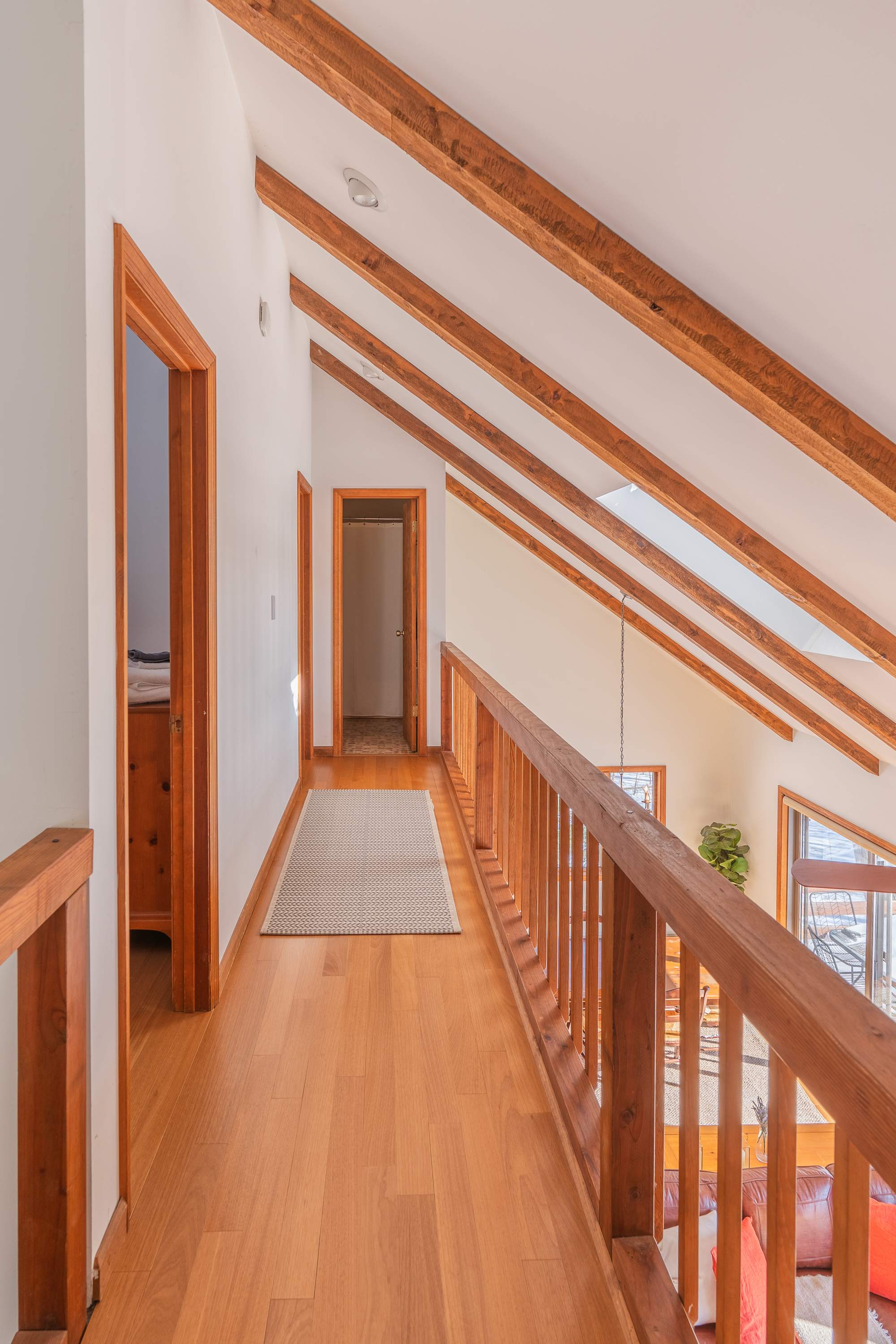 Upper-level hallway with hardwood floors and a wooden railing overlooking the living space below. Features exposed wooden beams on the vaulted ceiling and multiple doorways leading to other rooms.