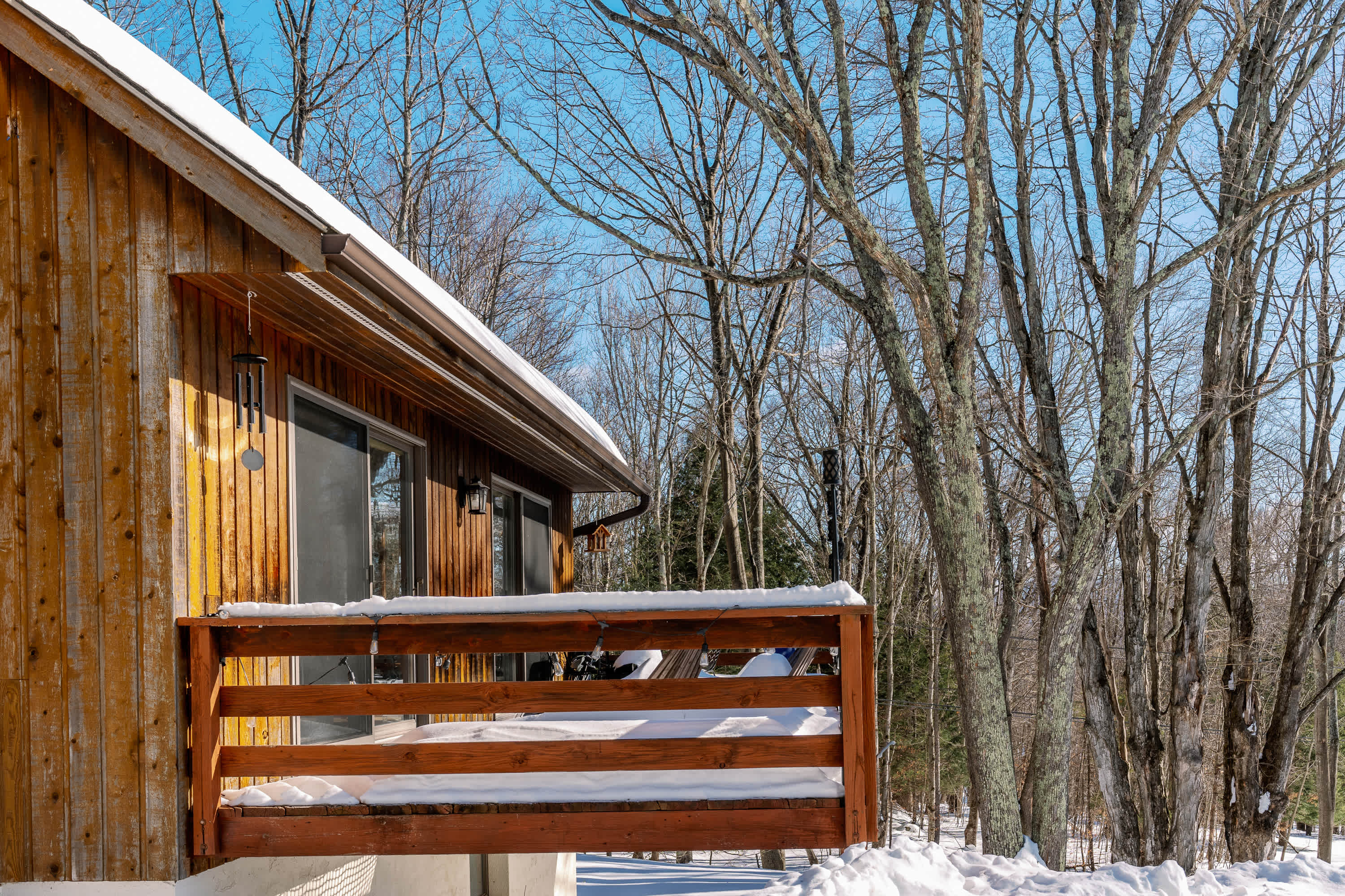 A wooden deck with snow-covered railings extends from a rustic wooden cabin, surrounded by bare winter trees under a clear blue sky. Sliding glass doors provide access to the interior, and snow piles up around the deck.