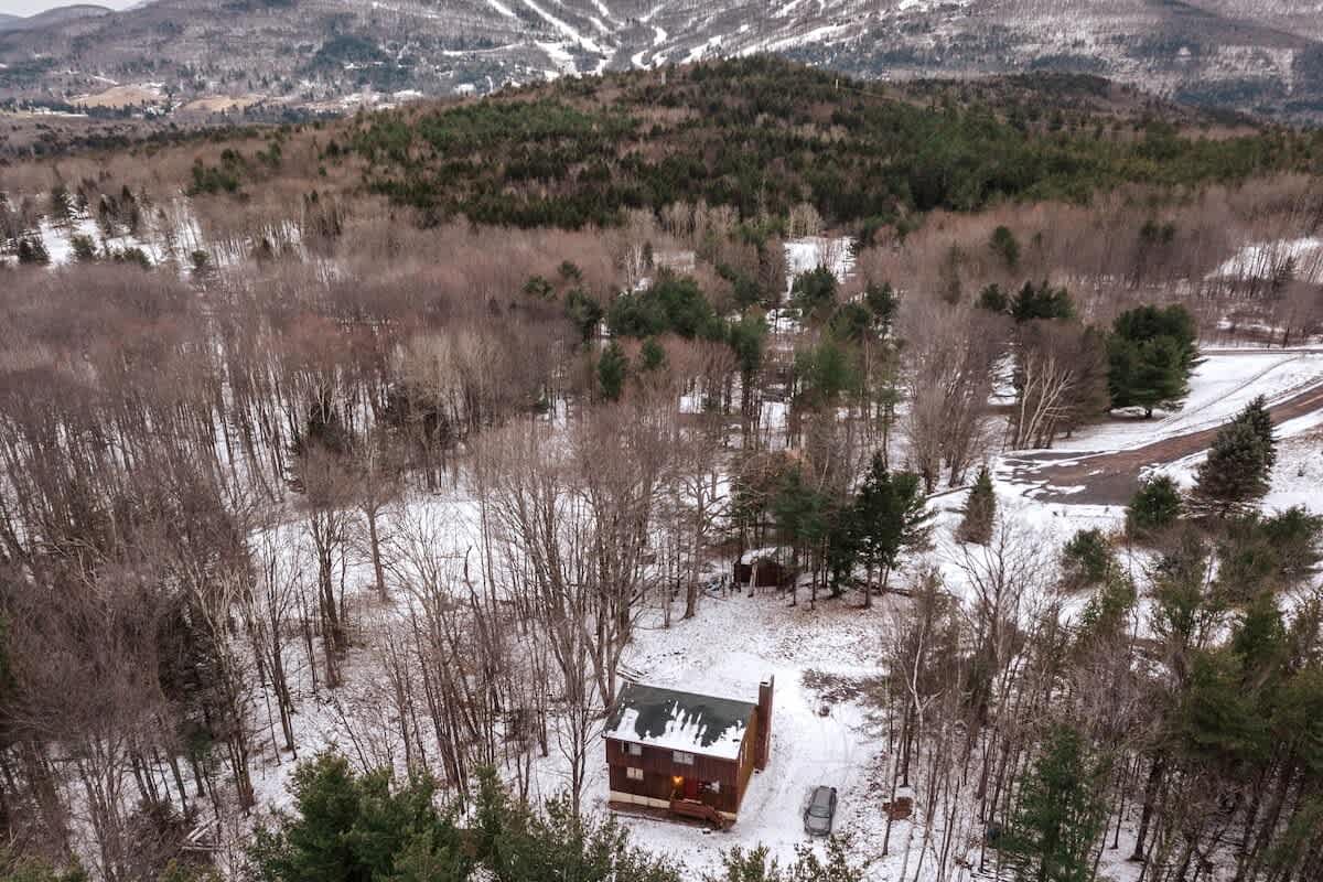 An aerial view of a brown cabin with a snow-covered roof, surrounded by bare trees and a snowy landscape. Distant mountains and ski trails are visible in the background under an overcast sky.