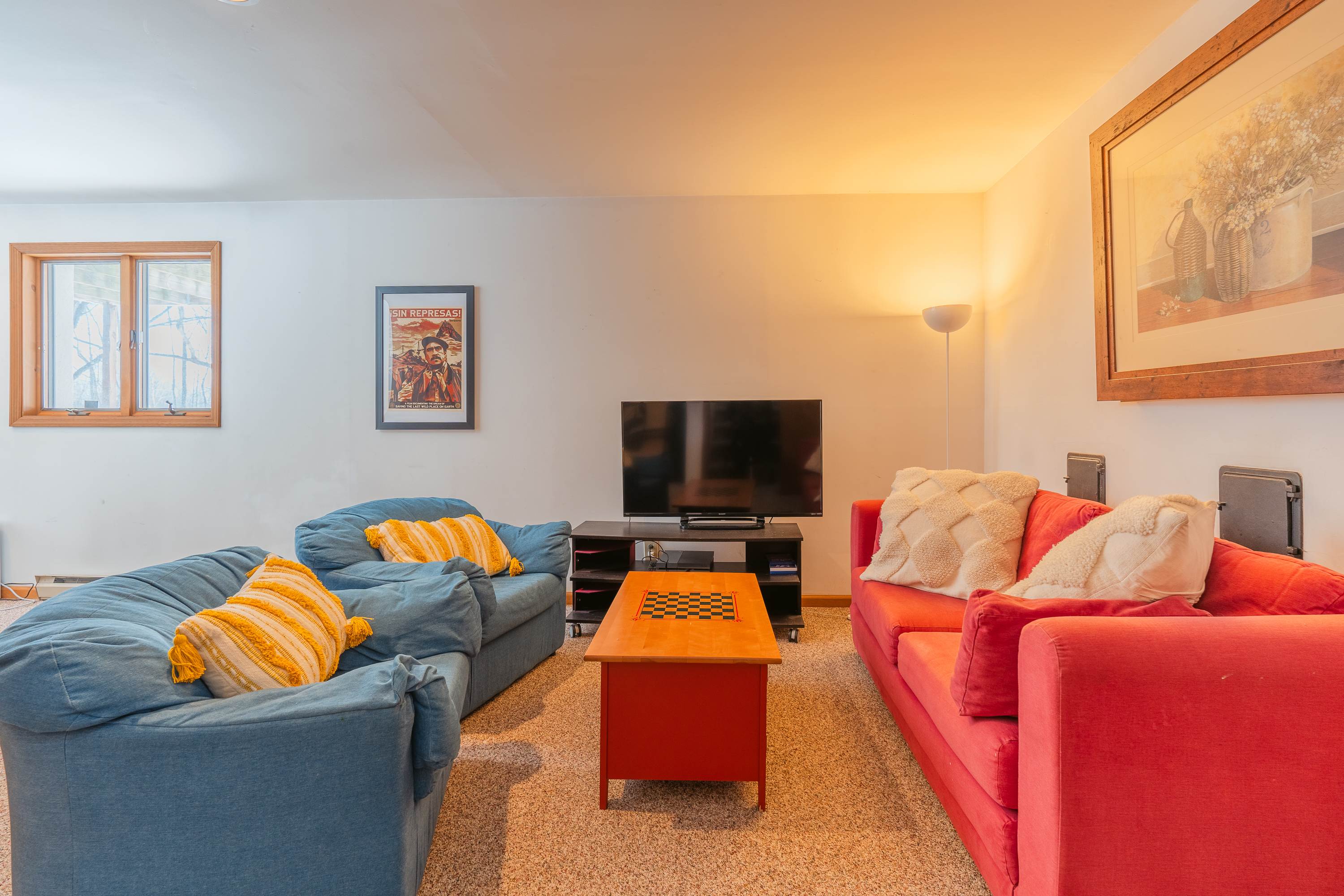 A vibrant living room featuring a blue sofa and a red sofa facing each other, with a red coffee table and a chessboard in the center. A flat-screen TV is mounted on a stand against the far wall, beneath a framed poster.