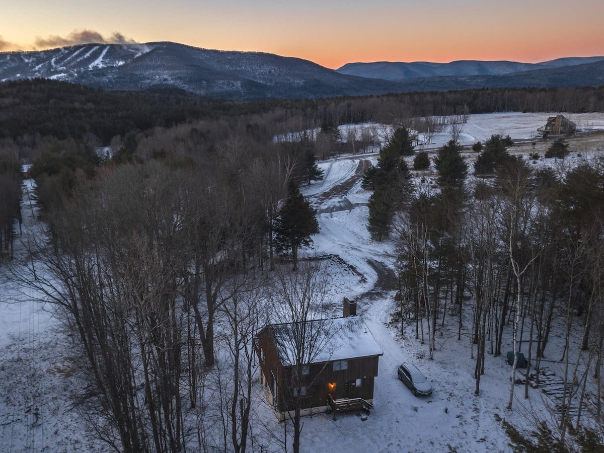 An aerial view of a rustic cabin nestled in a snow-covered, wooded landscape during sunset, with distant mountains and ski slopes visible under an orange-hued sky. A winding snowy driveway leads to the house.